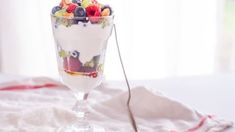 a glass filled with fruit and cereal on top of a white table next to a spoon