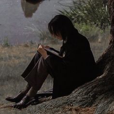 a person sitting on the ground reading a book next to a tree and water in the background