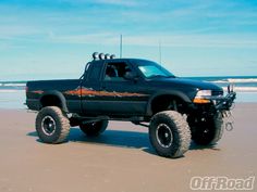 a large black truck parked on top of a sandy beach next to the ocean in front of blue sky