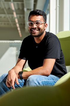 a man wearing glasses sitting on a green chair with his hand in his pocket and smiling at the camera