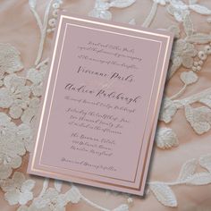 a pink and gold wedding card with white flowers on it's side, sitting on a lace covered bed sheet