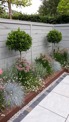 an outdoor garden with flowers and trees in the back ground, next to a white fence