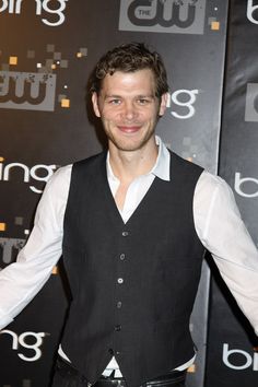 a man wearing a black vest and white shirt standing in front of a tv sign