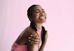 a woman with braids laughing and holding her hand on her chest while standing against a pink background