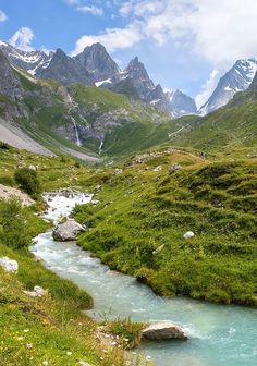 a river running through a lush green valley surrounded by mountains in the distance with rocks and grass on both sides