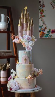 a three tiered cake with pink and blue frosting, flowers and castle decorations