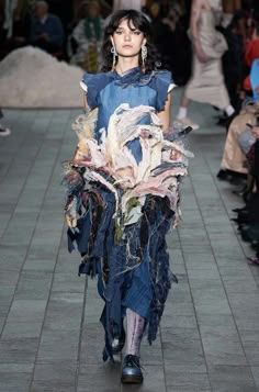 a woman walking down a runway wearing a blue dress with flowers on the skirt and large earrings