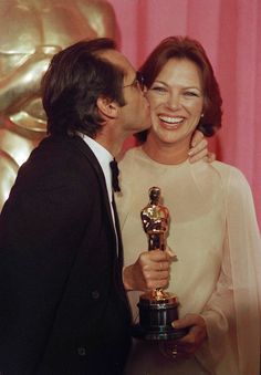 a man and woman kissing each other while holding an oscar award in front of a pink curtain