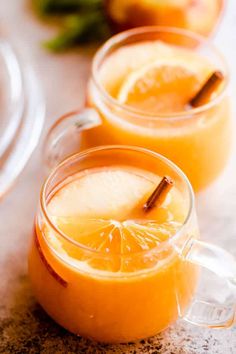 two mugs filled with orange juice and cinnamon stick garnished with an apple