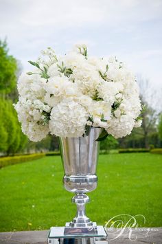 a silver vase filled with white flowers sitting on top of a wooden table next to a lush green field