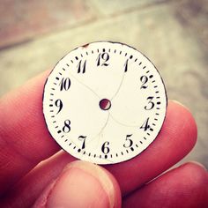 a hand holding a small white clock with black numbers on it's face in front of a brick wall