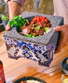 a person holding a bowl filled with food on top of a wooden table