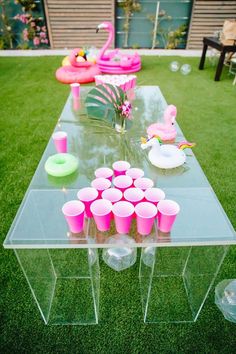 a clear table with pink cups and flamingos on it in the middle of grass