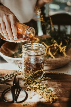 a person pouring tea into a glass jar on top of a wooden table next to scissors