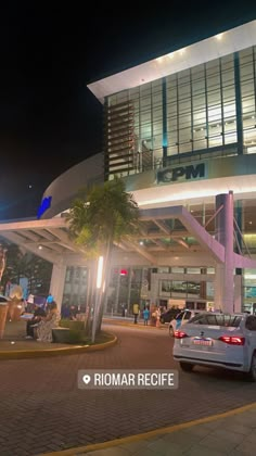 cars are parked in front of a building at night with palm trees and people walking on the sidewalk