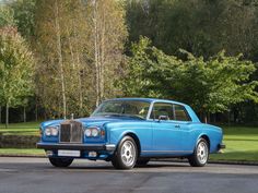 an old blue car is parked on the side of the road in front of some trees