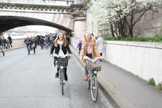 two women riding bikes down a street under an overpass