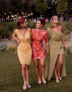 three women in dresses are standing together on the grass and smiling at each other while holding hands