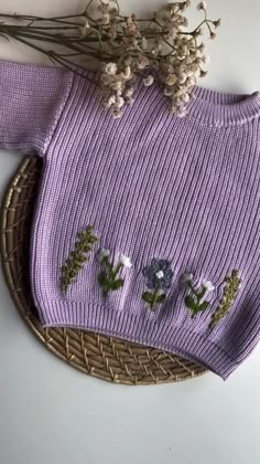 a purple sweater with flowers on it sitting next to a wicker basket filled with dried flowers