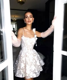 a woman in a short white dress is standing by a door and posing for the camera