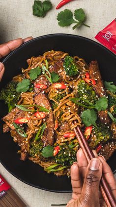 two hands holding chopsticks over a bowl of noodles with broccoli and beef