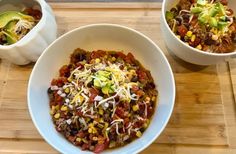 two bowls filled with chili, beans and cheese on top of a wooden cutting board