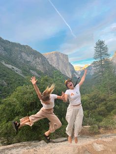 two people jumping up in the air with mountains and trees in the back ground behind them