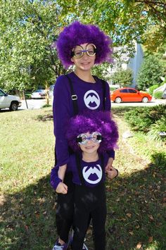 two children wearing purple wigs and glasses standing next to each other in the grass