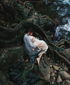 a woman holding a baby in her arms while sitting on the ground next to a river