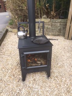 an outdoor wood stove with a kettle on the front and side burner, sitting in gravel