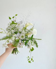 a hand holding a bouquet of white flowers and greenery in front of a wall