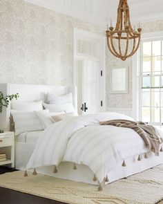 a white bed sitting in a bedroom next to a chandelier and two windows