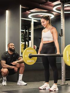 a man and woman in a gym setting, one is holding a barbell while the other squats down