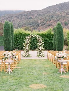 an outdoor ceremony setup with wooden chairs and white flowers on the back row, surrounded by greenery