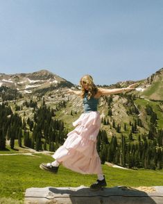 a woman in a long pink skirt is jumping on a bench with mountains in the background