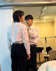 two young men standing next to each other in front of a mirror, one adjusting his tie