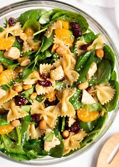 a glass bowl filled with pasta salad on top of a white table next to a wooden spoon