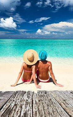 two people are sitting on the beach looking out at the blue water and white sand