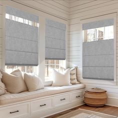 a window seat with pillows on it in front of two windows that have roman shades over them