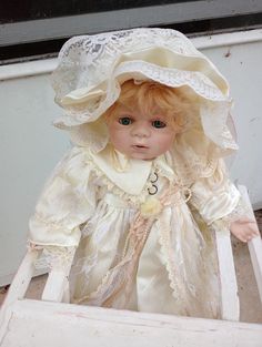 a small doll in a white dress and bonnet sitting on a wooden chair with a window behind it