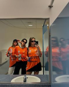 three women in orange shirts are taking a selfie with their cell phones while standing in front of a mirror