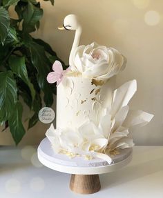 a white cake decorated with flowers and a swan on top is sitting on a table next to a potted plant