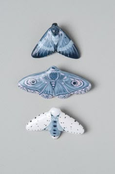 three ceramic figurines of moths on a gray background, one is blue and the other is white