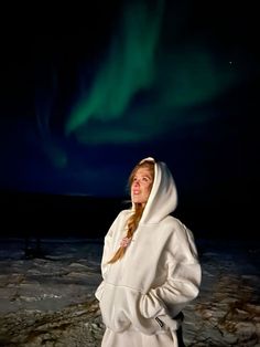 a woman standing in the snow wearing a hoodie and looking up at an aurora bore