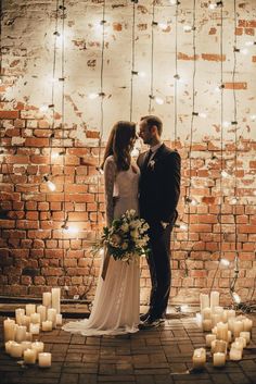 a bride and groom standing in front of a brick wall with candles