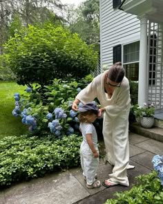 a woman and child standing in front of a house with blue flowers on the lawn