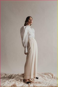 a woman standing on top of a white sheet covered bed wearing high heels and a long sleeved shirt