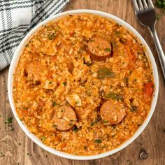 a white bowl filled with sausage and rice on top of a wooden table next to a fork