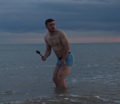 a shirtless man standing in the ocean holding a paddle and looking at the camera