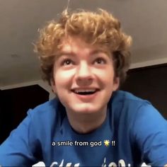 a young man with curly hair smiles at the camera while wearing a blue t - shirt